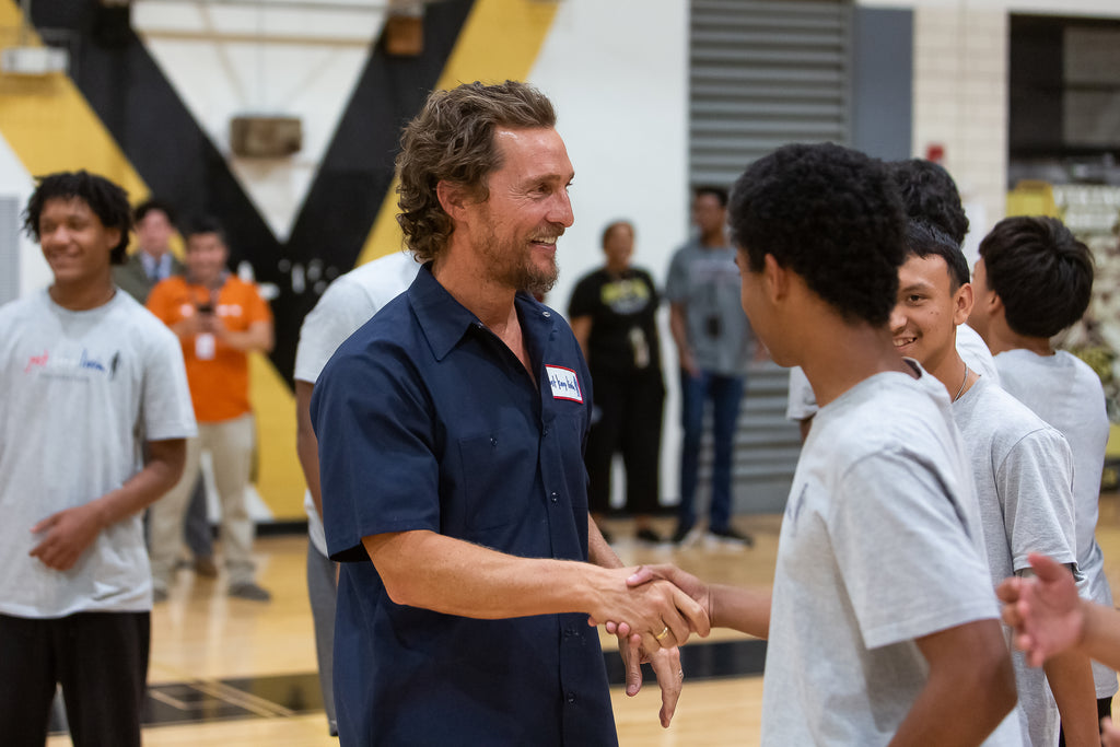 Matthew McConaughey in dark blue Just Keep Livin mechanic shirt shaking hands with a young boy in a gymnasium setting