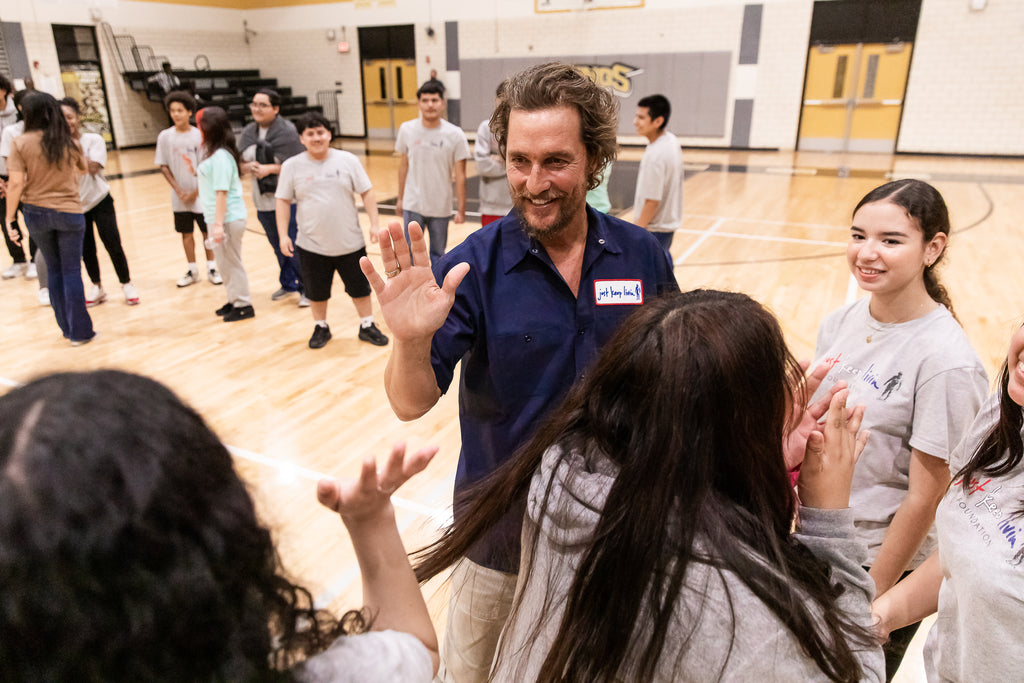 Matthew McConaughey giving a high five to a just keep livin student at an event in a school gym.  Matthew is wearing the dark blue mechanic shirt the foundation sells. 