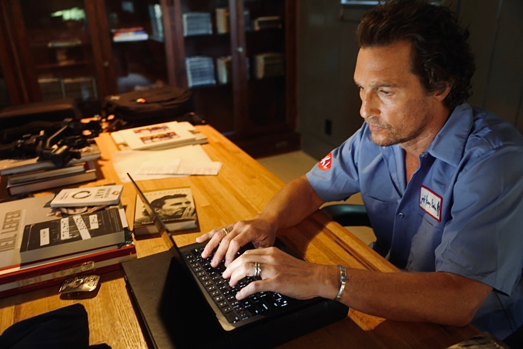 Man in a light blue mechanic shirt with a just keep livin logo sitting at a desk using a laptop.