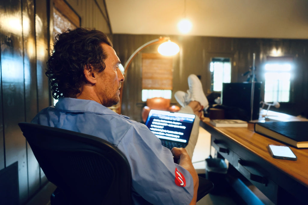Man sitting at a desk using a laptop in a dimly lit room.  He is wearing the mechanic shirt being sold by the store. The shirt is light blue. 
