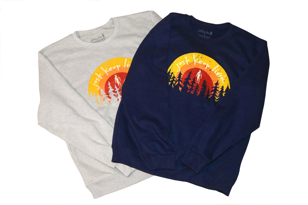 Two crewneck sweatshirts, one in heather gray and one in navy, both with a screenprinted design of a sunset over a forest and the 'just keep livin' logo.