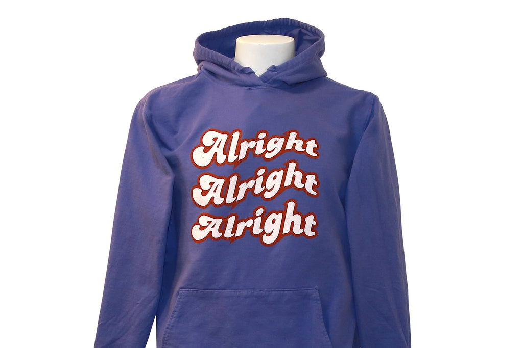 A midweight pullover hoodie in blue with the text 'Alright Alright Alright' printed in a repeating pattern across the front with white text with red outline.